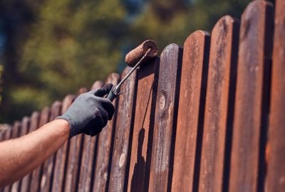 man-protective-gloves-is-painting-wooden-fence-bright-summer-day-min